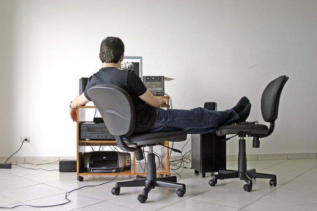 Muž v bílé místnosti se stolem a počítačem, nohy má na vedlejší židli, pracuje